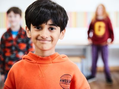 A young boy with black hair and wearing a bright orange jumper smiles at the camera